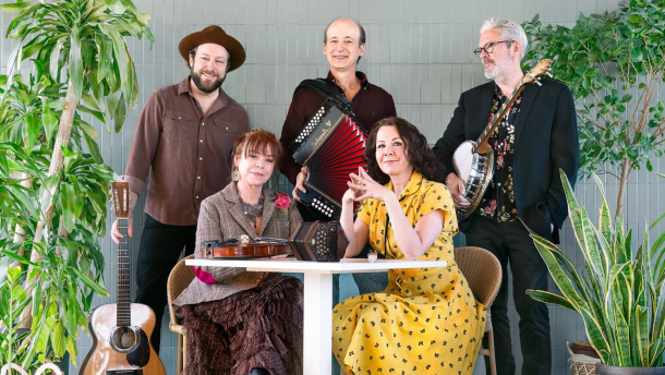 Five musicians smile at the camera in a room surrounded by verdant plants.