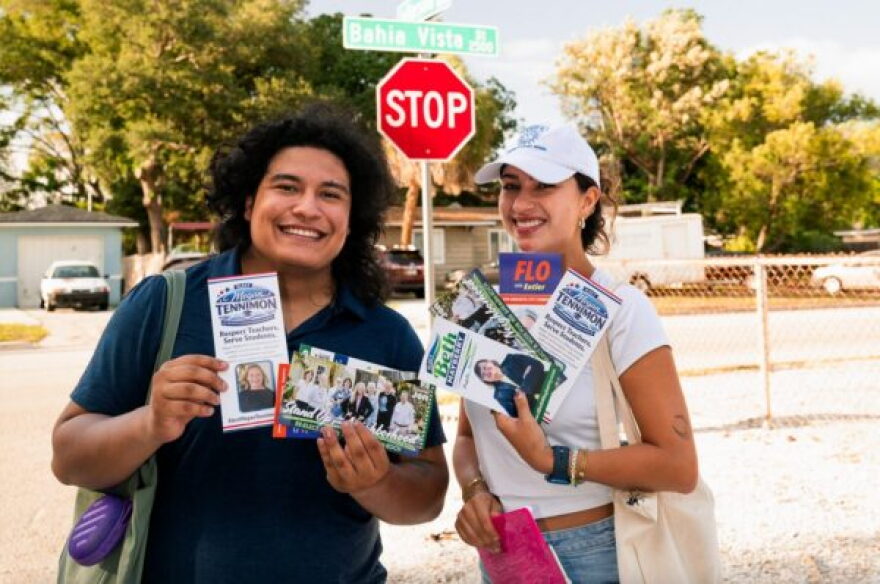 Two people outside holding campaign materials with a stop sign and houses behind them 