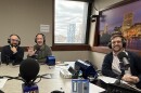 Three men wearing headphones sit in front of microphones at a table in a radio talk studio: the man on the left is bald, has a dark beard and is wearing glasses, a white button down shirt and navy sweater; the man in the center has short brown hair and is wearing a green sweatshirt; the man on the right has short brown hair and is wearing glasses, a white button down shirt and navy cardigan.