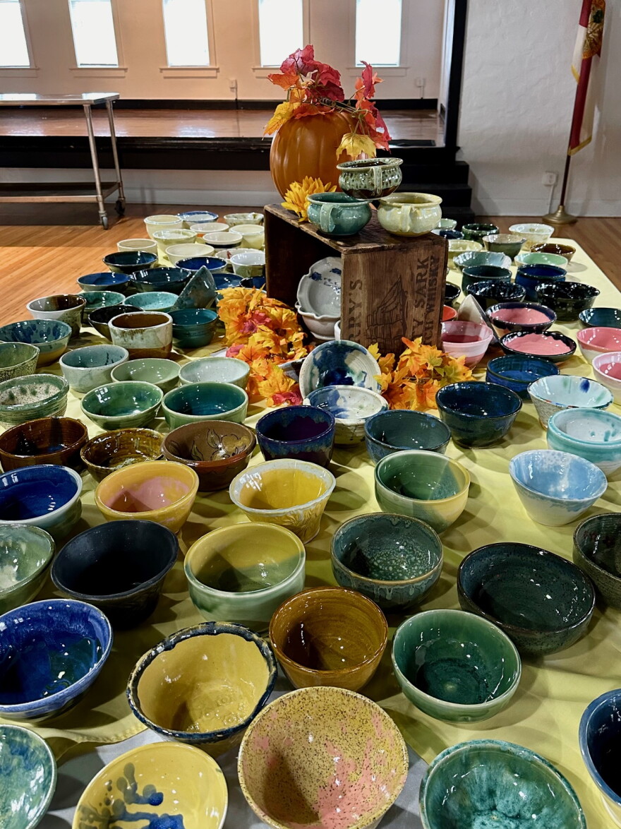 Handmade ceramic bowls in blues, greens, yellows, browns and pinks cover a long table with a yellow tablecloth in a wood-floored hall. At the center, a wooden crate holds two handled soup bowls, with an orange pumpkin of autumn leaves and a flag in the background.