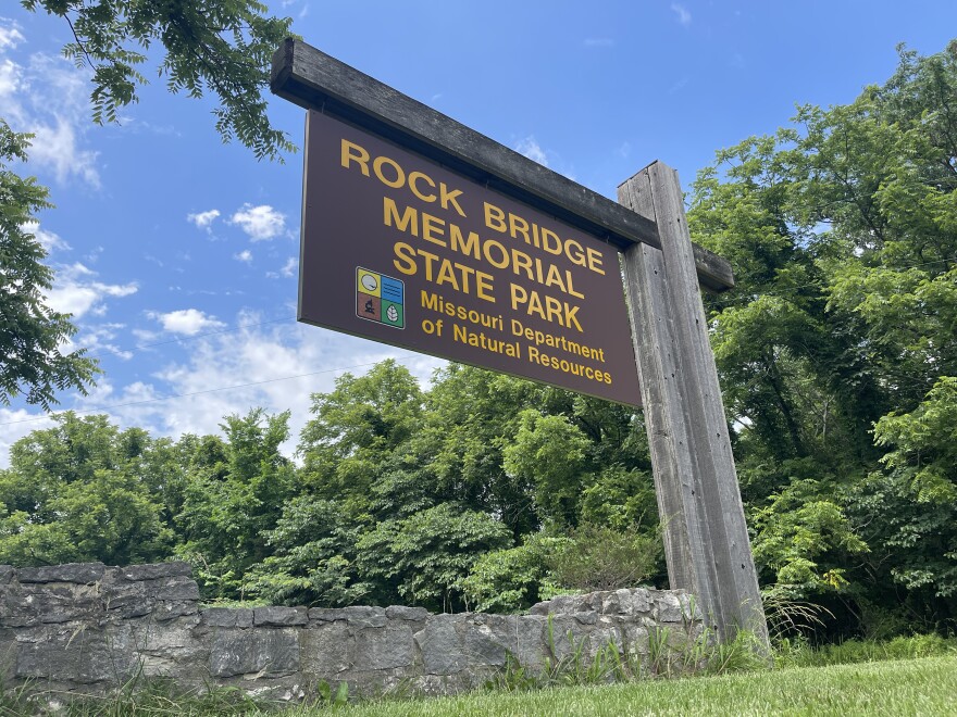 A large brown sign with yellow lettering says, "Rock Bridge Memorial State Park, Missouri Department of Natural Resources." In the backdrop is a bright green forest.