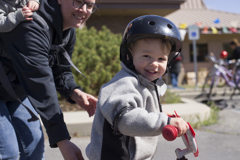 A child with a helmet on smiles at a camera as he sits on a bike.