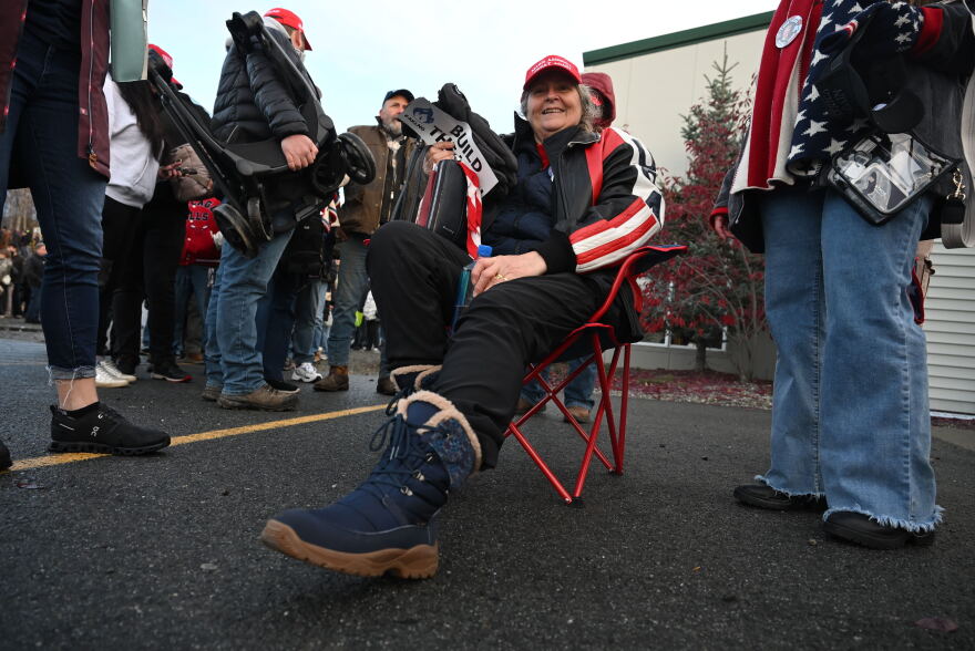 Juneau resident Cindy Fuller sits in a chair with an American flag print near the shuttle to get to President Donald Trump's midterm rally in Wasilla.