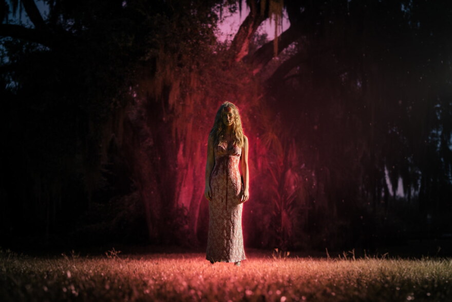 Girl in long dress stands outside in grass in front of large trees as she's covered in fake blood. 