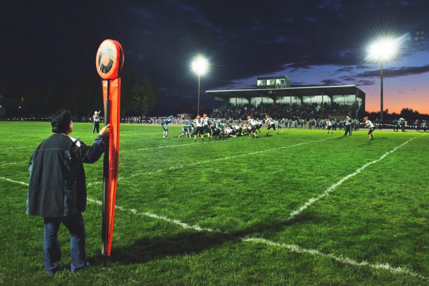 A person holding an orange down-marker on the side of a football field with a football game in the background