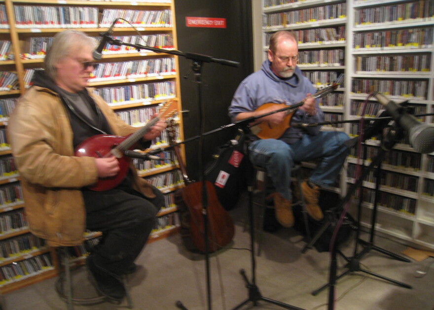 Two men seated on stools in a radio studio playing mandolin