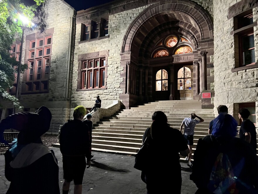 A group of people wearing black face a stone building, lit at night. 