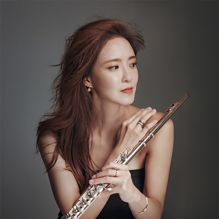 A young woman in a sleeveless black gown, and holding a silver flute looks away from the camera.