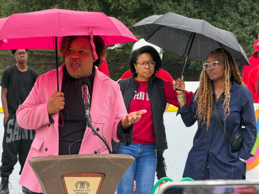 A woman in a pink jacket holds an umbrella and speaks behind a podium.