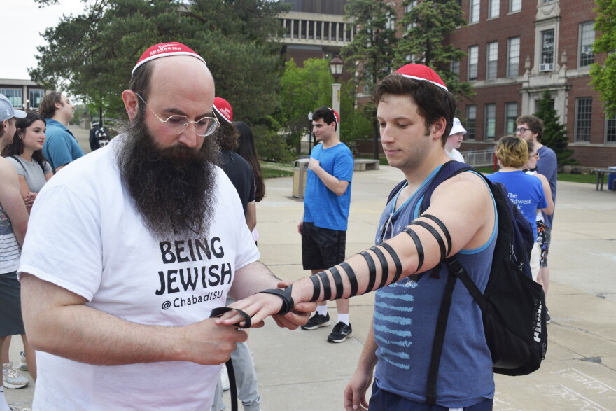 Bearded man wearing a red kippah wraps a tefillin around the left arm of a college student in an outdoor plaza area