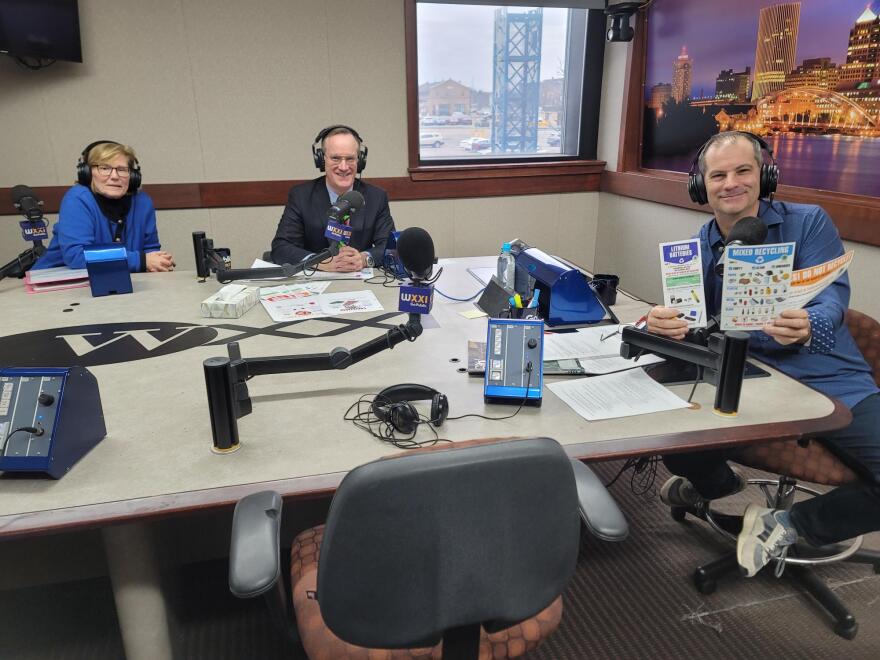 Three people wearing headphones sit at a table in a radio talk studio: a woman at left has short blonde hair and is wearing glasses, a blue sweater, and black shirt; a man at center has short brown hair and is wearing glasses, a navy blazer, a light blue button-down shirt, and a green and red patterned tie; a man at right has short dark hair, is wearing a blue button-down shirt and jeans and is holding several brochures showing recycling instructions.