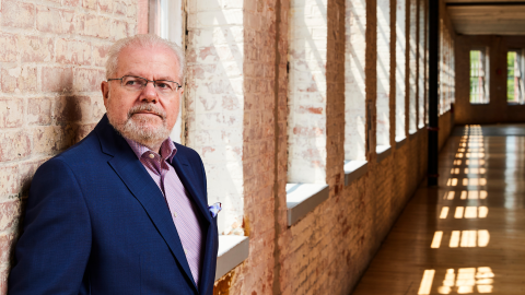 Emanuel Ax wears glasses, a navy suit, and a pink button down. He stands in a stone building with sun pouring in through it's windows.