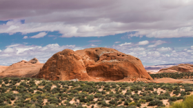 A large, slightly rounded red rock outcropping in a desert landscape.
