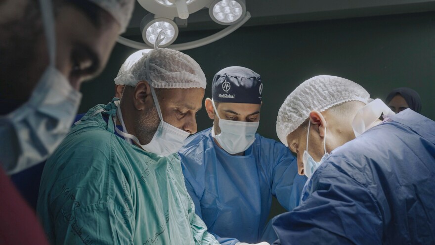 Four medical practitioners in different-colored scrubs (red, light teal, and light blue), face masks, and hair coverings lean over an offscreen patient while conducting surgery on them.