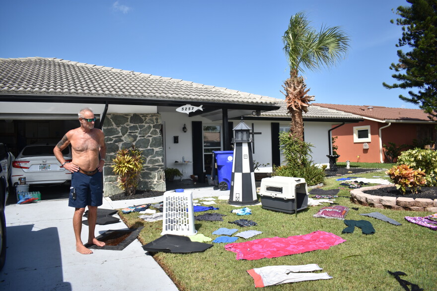 A man without a shirt stands in his driveway with his hands on his hips surveying the array of items laid out to dry on his front lawn. 