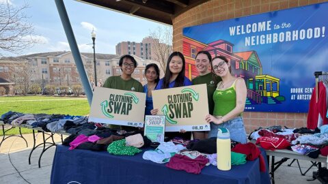 A group of five students stand behind a table covered in stacks of clothes. They hold signs that say, 'Clothing Swap.'