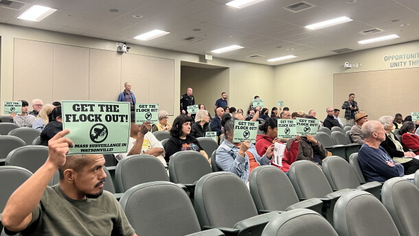 An auditorium is filled with people, many holding signs that read "Get the Flock Out."