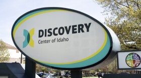 The outside oval sign of the Discovery Center of Idaho near its parking lot. It is white, with black lettering and yellow/turquoise highlights. 