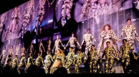 A line of women in gold outfits dance on a large stage during a concert in a stadium atmosphere.
