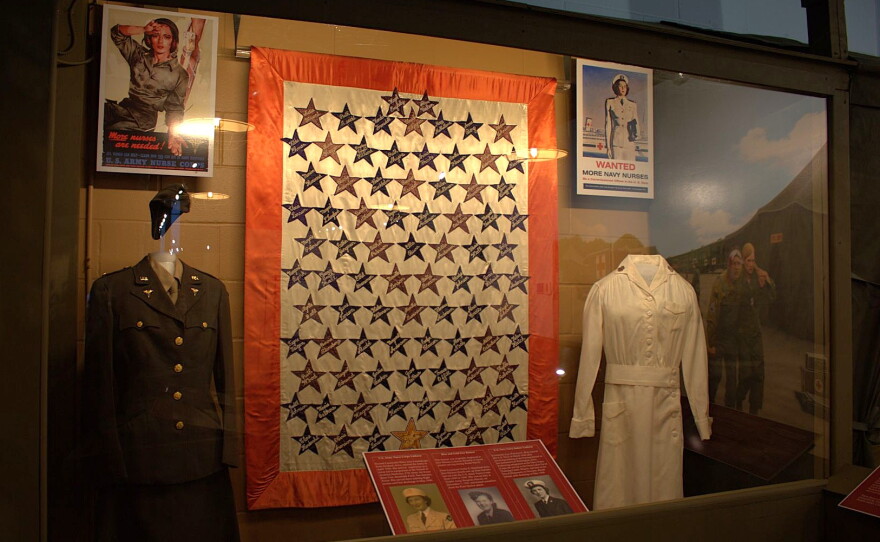 An army and navy nurse uniform on display in front of corresponding recruitment posters, and a tapestry with the names of many Iowa nurses who served.