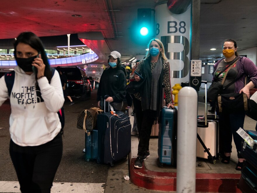 Travelers wearing protective face masks wait for a shuttle to arrive at the Los Angeles International Airport in Los Angeles, Tuesday, Nov. 30, 2021.