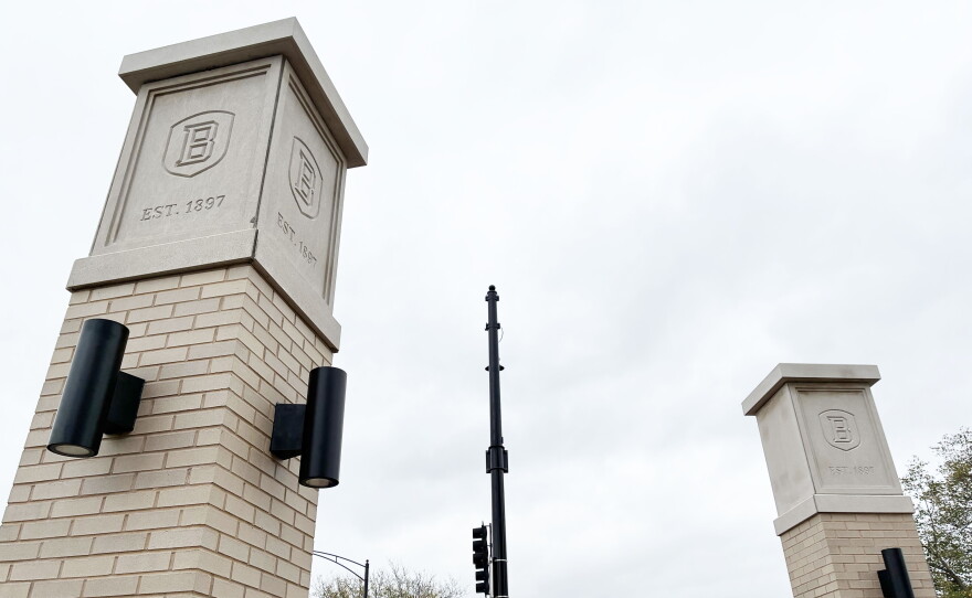 Two light-colored brick pillars topped with stone markers engraved with a “B” and “EST. 1897,” standing on either side of a traffic light pole under a cloudy sky.