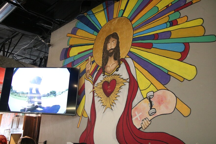A giant colorful mural shows Jesus in a white robe and red sash holding two fingers up with one hand and holding a pink skateboard with a cross on it in the other hand. There are yellow, red and blue lines coming out from a gold circle around the back of his head.