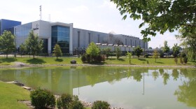 A photo of a data center from the street nearby. Behind a fence and across a green pond, fans on the roof of the center exhaust a dark smoke.