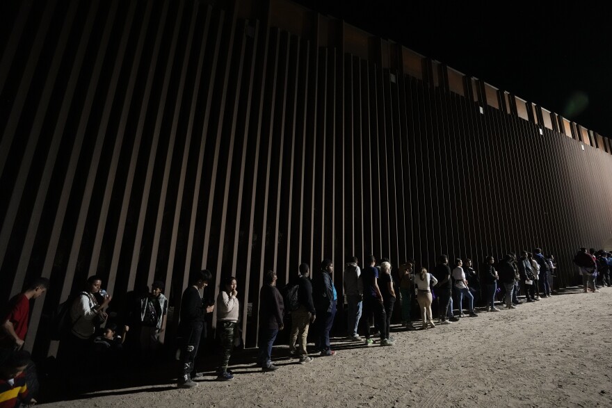 People line up against a border wall as they wait to apply for asylum after crossing the border from Mexico, July 11, 2023, near Yuma, Ariz.