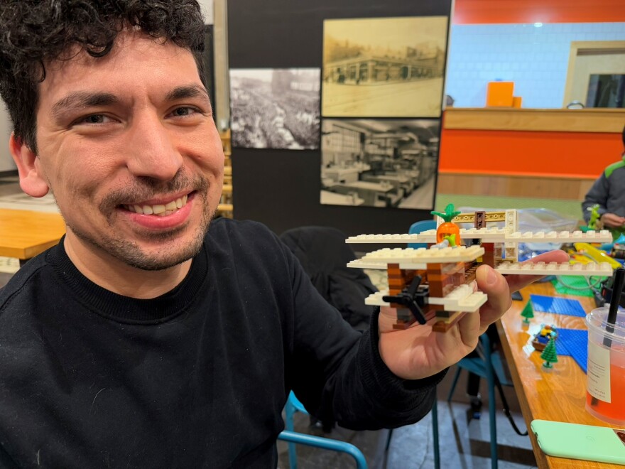 A man with brown hair and a short beard is wearing a black long-sleeved T-shirt and holds up an airplane made of Legos.