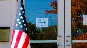 A sign on a glass door reads, "voting today" with an arrow. An American flag is in the foreground. 