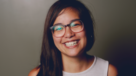 Danni Quintos with long dark hair smiling at camera wearing a tank top and glasses with a tattoo on her right upper arm.