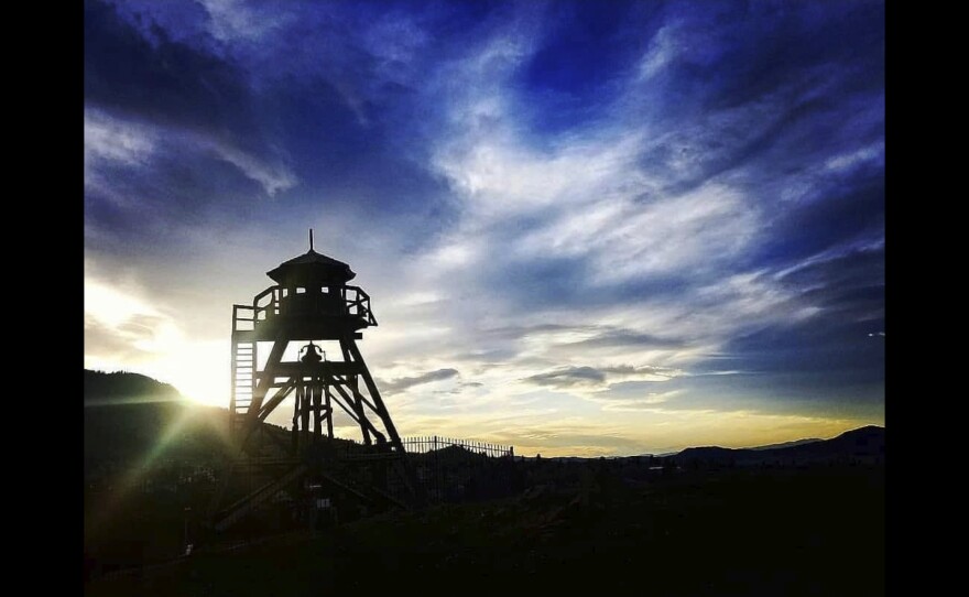 A fire lookout tower stands on the horizon in front of a setting sun and clouds of orange and blue.
