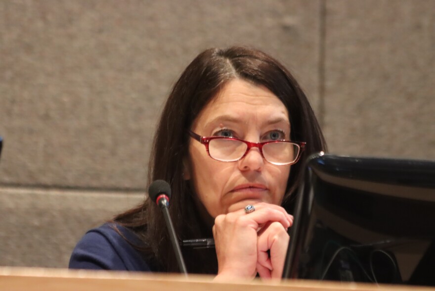 A white woman with black hair and red glasses rests her chin on her hands in front fo a microphone