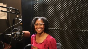 A smiling woman in a pink blouse sits in a recording studio holding headphones beside a microphone, with soundproof foam panels behind her.