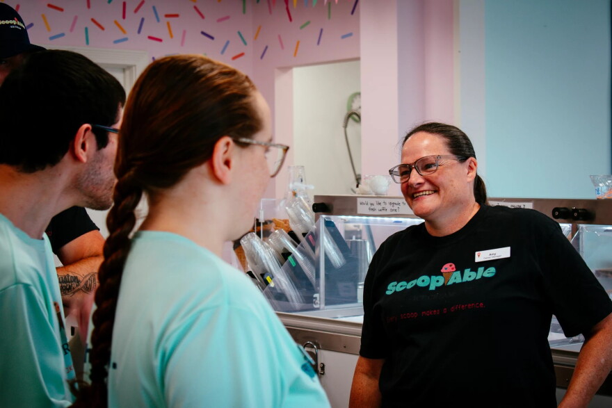 a woman in a black tshirt stands smiling before a group of people in teal shirts, who are pictured from behind