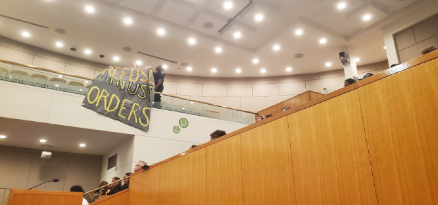 At a CMS school board meeting, an attendee unfurls a banner to protest ICE at schools.
