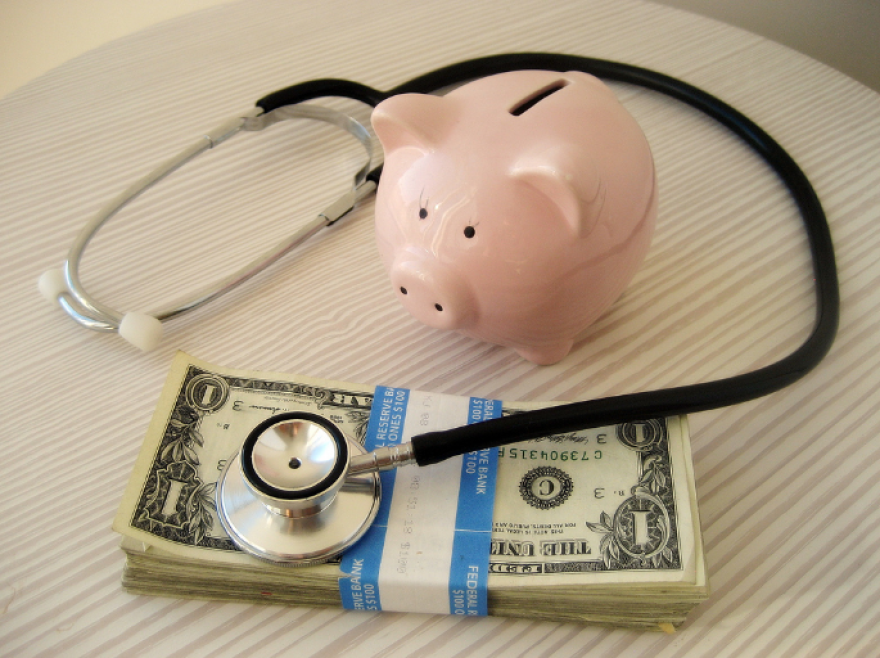 a piggy bank, a stack of one dollar bills, and a stethoscope sit on a woodgrain table 