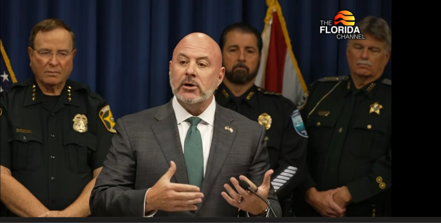 Un hombre calvo con traje habla desde un podio, flanqueado por agentes de la ley.