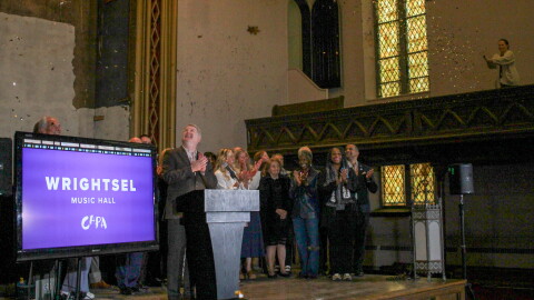 People on a stage clap as confetti flutters through the air in an old church. A tall, thin stained glass window glows in one corner. A TV screen displays the name "Wrightsel Music Hall / CAPA" 