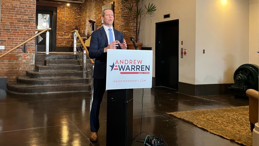 Man in suit speaks at a podium with the name 'Andrew Warren' on it. A short stairwell and an elevator door are behind him. 