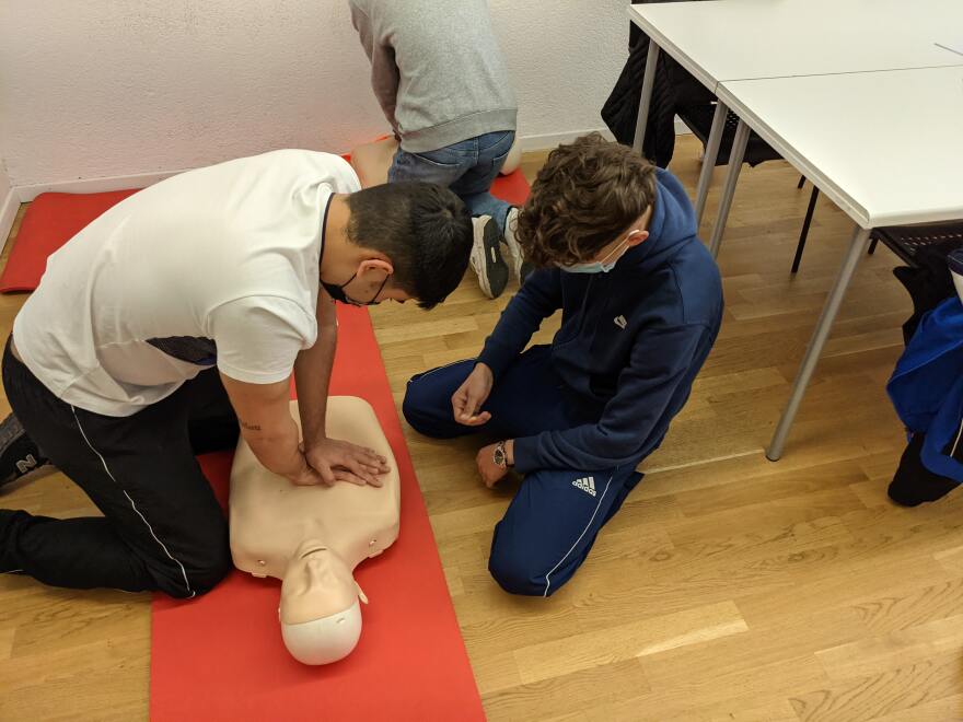 a man in a white shirt and black pants wears a mask and practices administering cpr on a dummy laying on a red mat