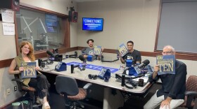 Four smiling people holding copies of CITY Magazine as they sit around a table in a radio talk studio: a woman front left has long wavy red hair and is wearing a green top, black pants, and white sandals; a man back left has short curly brown hair and is wearing a gold chair and a blue graphic t-shirt; a man back center has short dark hair and is wearing glasses, a gold chain, and a black graphic t-shirt; a man at right has very short dark hair and is wearing a black button-down shirt, tan linen pants, and grey and blue sneakers