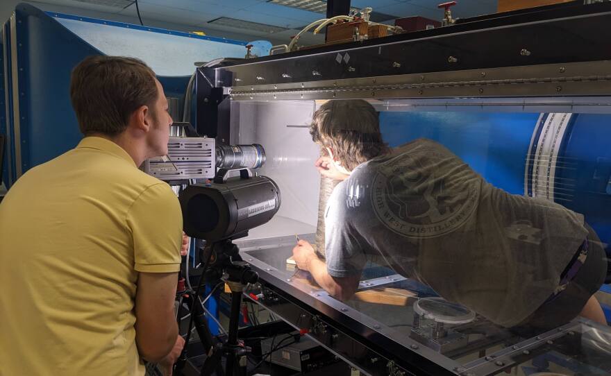Two graduate student researchers Rhett Parry (left) and Mike Wayman (right) calibrate the wind tunnel, a large plastic and metal structure within a lab with a tree trunk in the middle