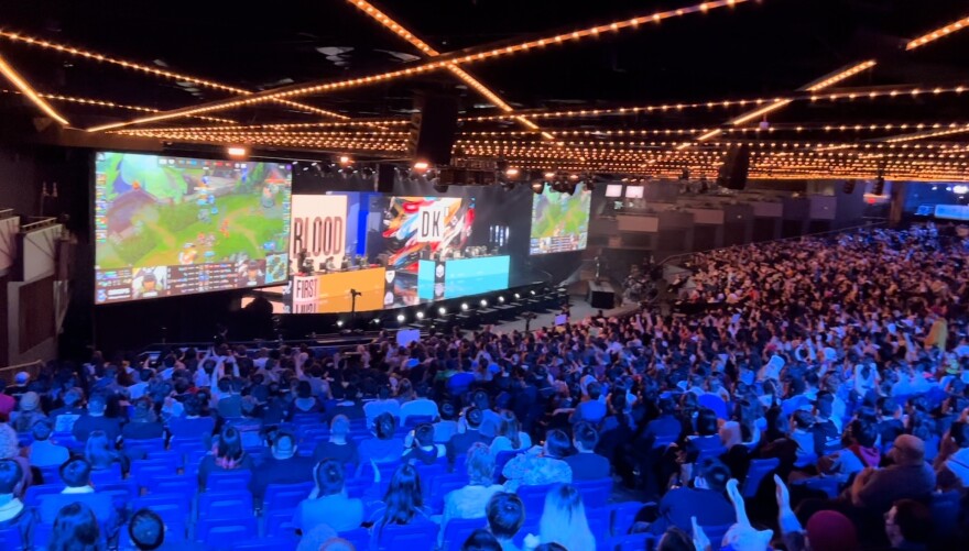 A large crowd in a theater looks at a stage with two tables and large television displays.