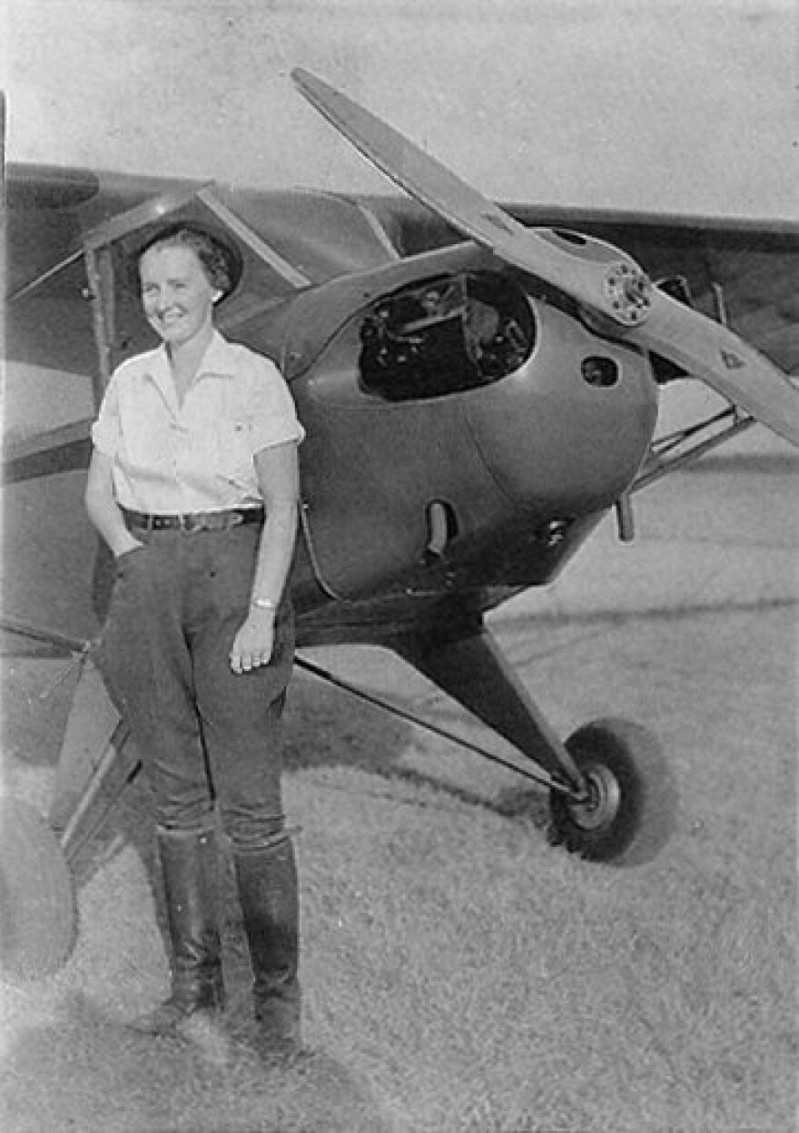 A black-and-white photo shows a woman standing beside a small propeller airplane.