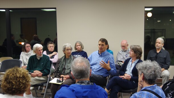 Attendees sit in an oval of fold out chairs, with only a portion shown in the photo. A man in a light blue button up gestures with his hands as he speaks to the crowd, who all look at him. 