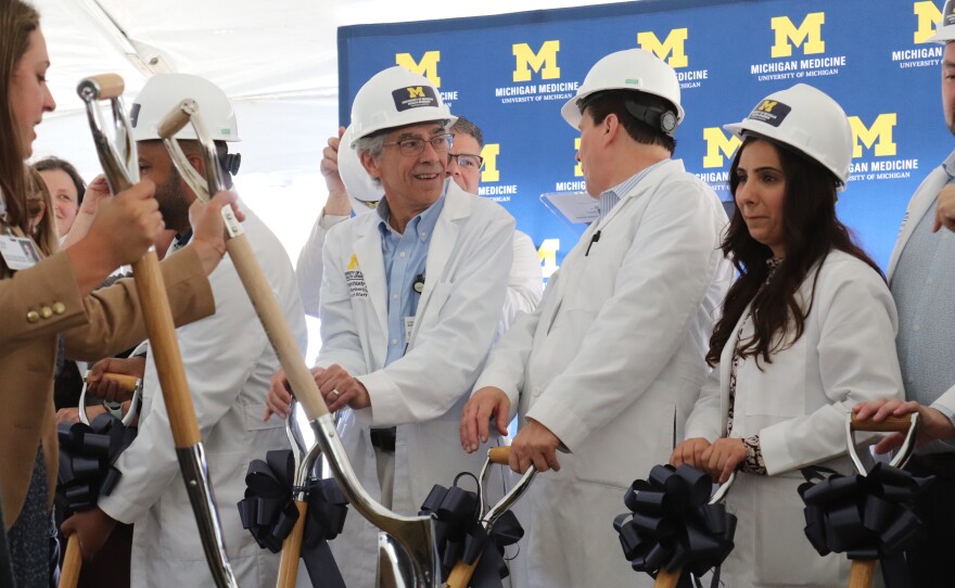 People in lab coats and hard hats hold shovels at a groundbreaking event, with a University of Michigan Medicine backdrop behind them.