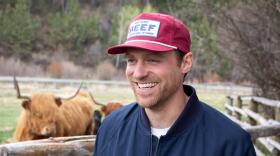 A man in a red "BEEF" ball cap smiles in front of a pair of highland cows in a corral.