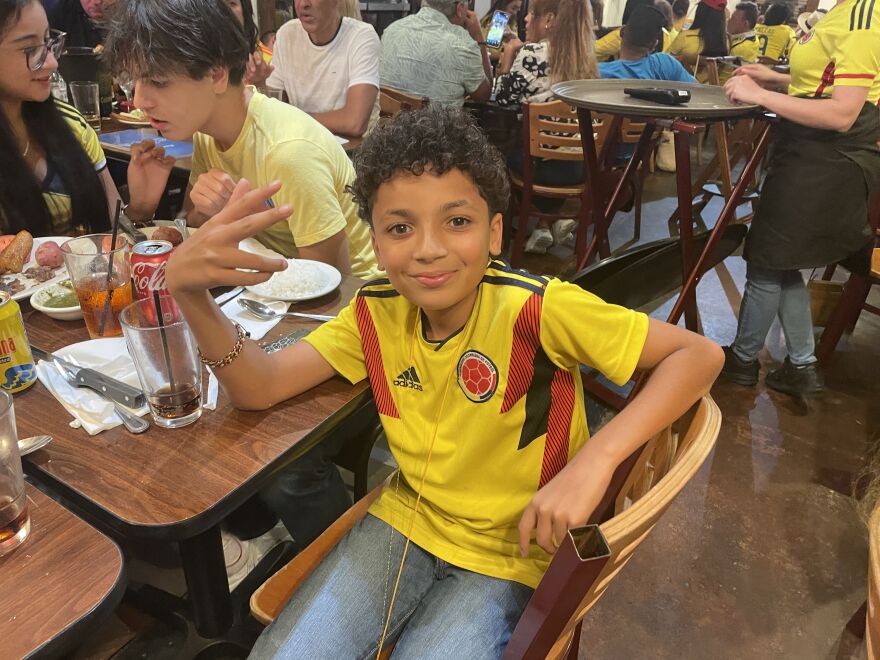 A boy wears a Venezuelan soccer jerset and light blue jeans. He's looking at the camera and smiling.